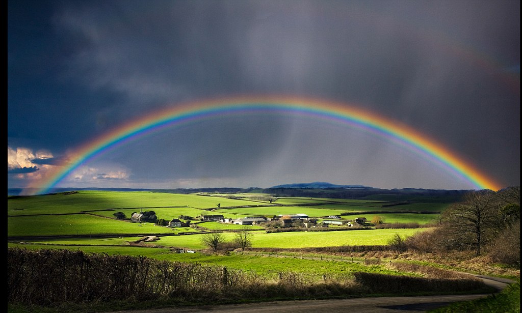 A full rainbow over a farm illumnated by sunlight amongst a rain storm