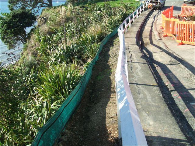 A roadside on a coastline and a green wind-cloth fence blocking a large amount of soil from flowing further down the coastside.